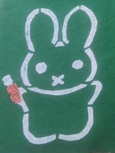 A stencil of a green bunny in a white outline, holding an orange carrot.